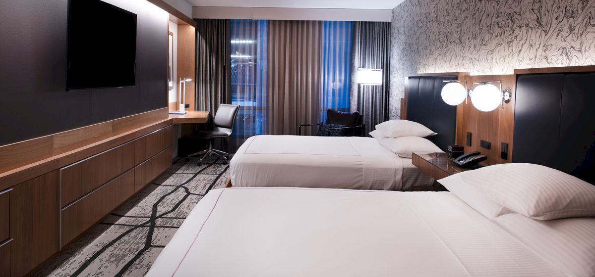 The image shows a modern hotel room with two beds, a large TV, desk, and window with curtains.
