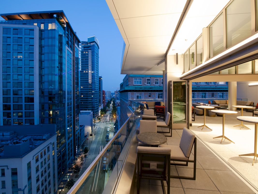 A cityscape view from a high-rise balcony, featuring modern buildings and a well-lit terrace with seating during twilight.