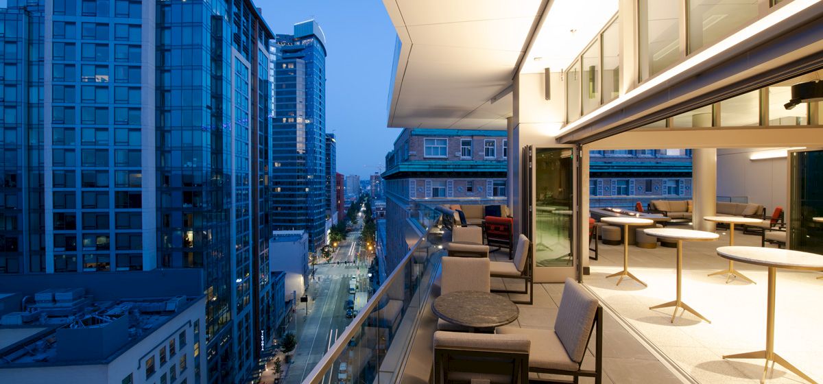 A cityscape view from a high-rise balcony, featuring modern buildings and a well-lit terrace with seating during twilight.