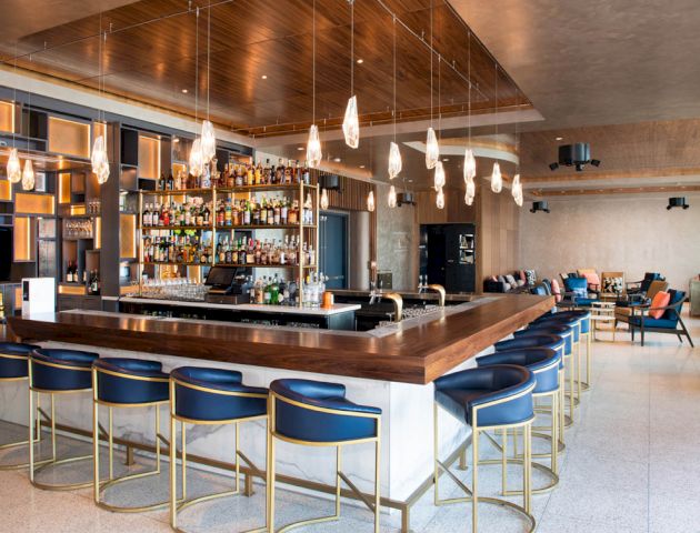 The image shows a sleek bar with blue stools, modern lighting, and a well-stocked shelf, creating a stylish and inviting atmosphere.