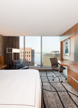 The image shows a modern hotel room with a bed, large TV, desk, and city view from a window. The decor is sleek with wooden elements.