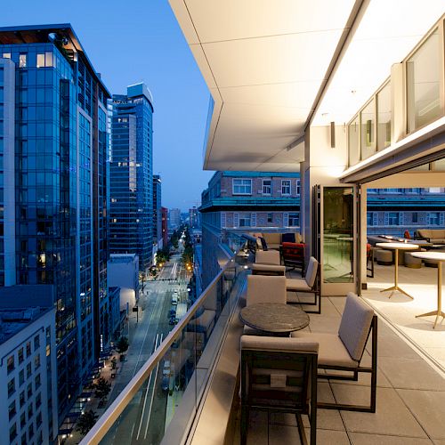 A modern rooftop terrace overlooks an urban street with high-rise buildings under the evening sky, featuring seating areas and tables.