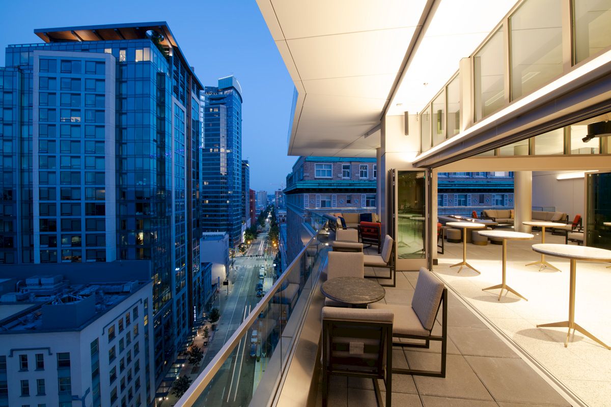 A modern city balcony during twilight with seating and tables overlooks tall buildings and a street.