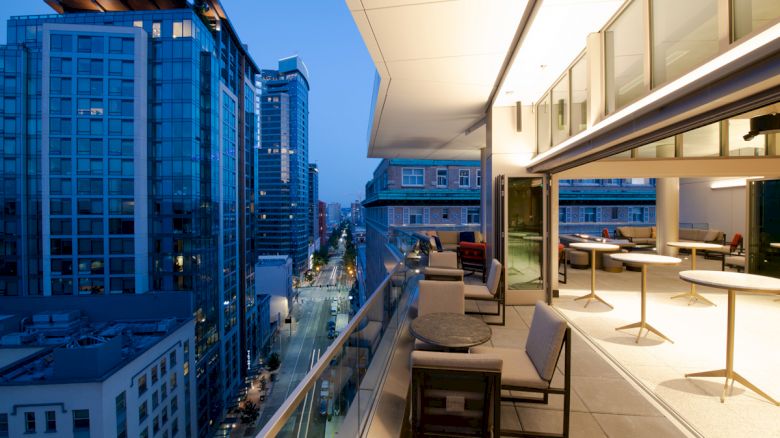 A modern city balcony during twilight with seating and tables overlooks tall buildings and a street.