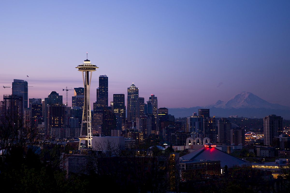 The image shows the Seattle skyline at dusk, featuring the Space Needle with a mountain in the background.