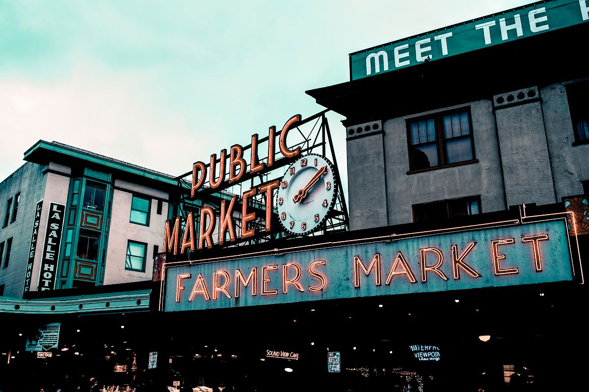 A public market with neon signs, including a clock and "FARMERS MARKET," on buildings in an urban setting.