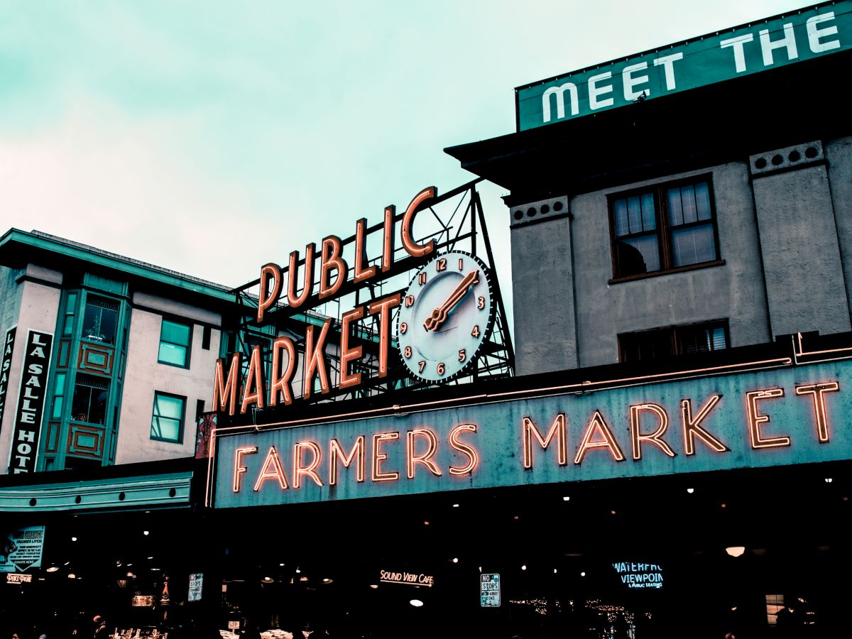 A public market with neon signs, including a clock and "FARMERS MARKET," on buildings in an urban setting.