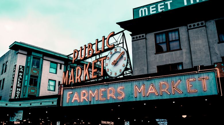 A public market with neon signs, including a clock and "FARMERS MARKET," on buildings in an urban setting.