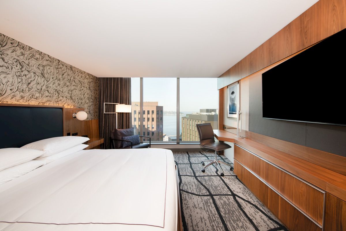 A modern hotel room with a large bed, desk, chair, and a flat-screen TV. Large windows offer a city view with natural light.