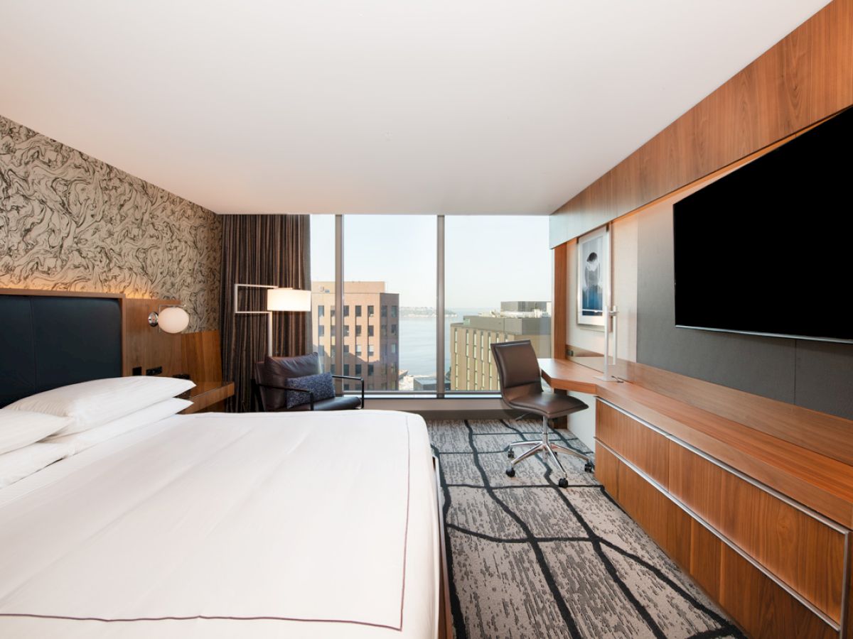 A modern hotel room with a large bed, desk, chair, and a flat-screen TV. Large windows offer a city view with natural light.