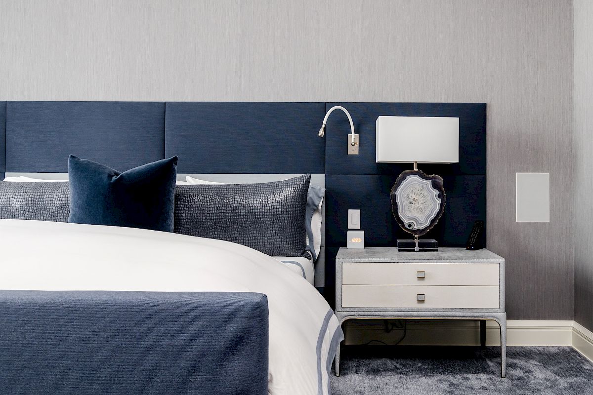 A modern bedroom with a blue and gray color scheme features a bed, side table, lamp, pillow, and decorative items.