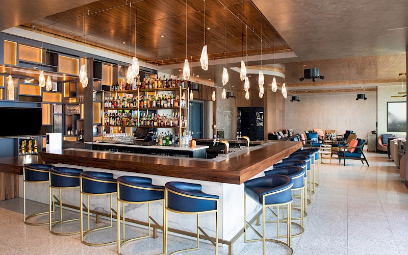 The image shows a stylish bar interior with blue barstools, hanging lights, and a well-stocked counter, creating a cozy atmosphere.