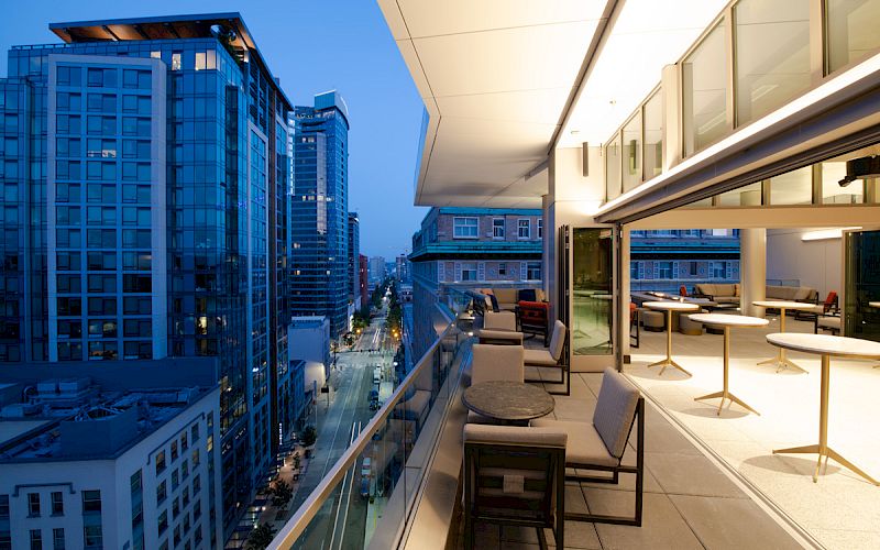 A modern rooftop terrace with seating overlooks a cityscape featuring tall buildings at dusk.