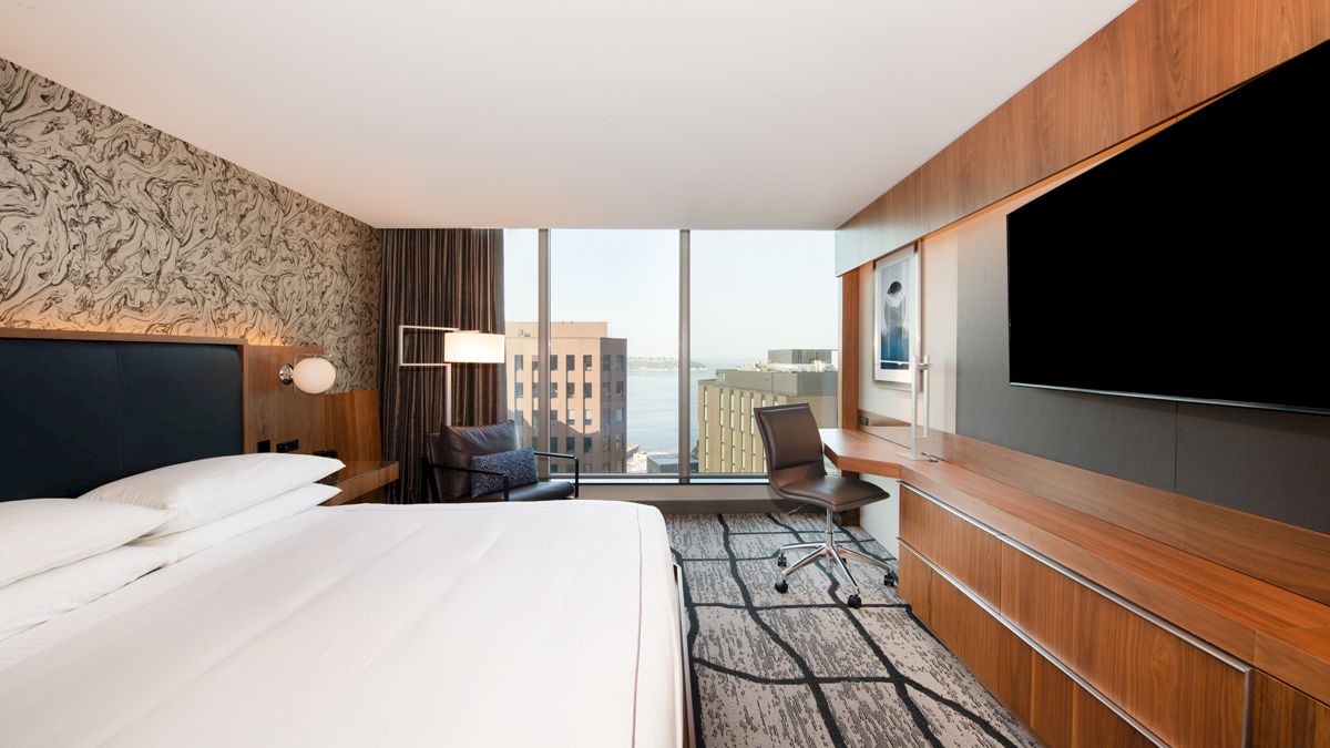 A modern hotel room with a large bed, desk, and TV, featuring a city view through a large window.